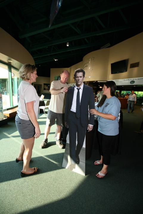People gather around a life-sized man cutout in a well-lit indoor space with green flooring.