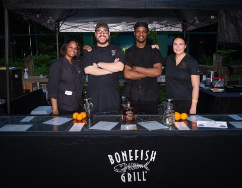 Four smiling chefs in black uniforms stand behind a Bonefish Grill counter, arms crossed, under a tented outdoor setup.
